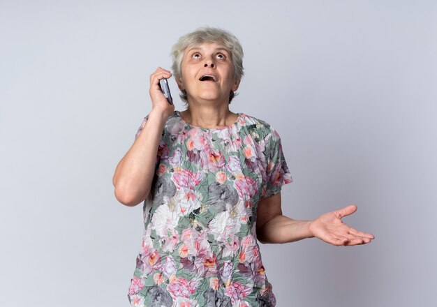 Excité femme âgée parle au téléphone et lève les yeux isolé sur mur blanc