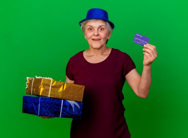 Excité femme âgée portant chapeau de fête détient des coffrets cadeaux et carte de crédit sur vert