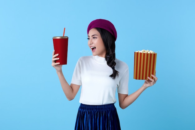 Excité femme asiatique regardant un film film tenant du pop-corn et une tasse de soda sur bleu vif.