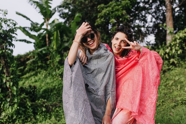 Excité femme brune en imperméable rose s'amusant avec son meilleur ami. Photo extérieure d'adorables sœurs passant du temps dans une forêt exotique.