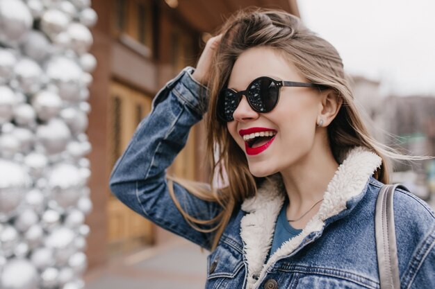Excité femme caucasienne dans des lunettes de soleil à la mode en détournant les yeux avec un sourire surpris. Plan extérieur d'un gitl agréable en veste en jean riant le week-end.