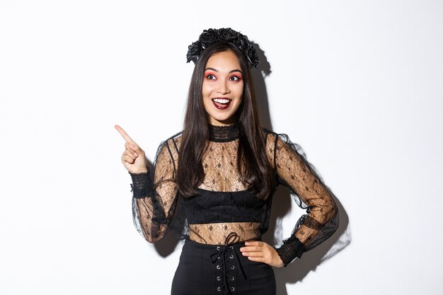 Excité heureux femme asiatique en robe de dentelle noire et guirlande à la stupéfaction dans le coin supérieur gauche, pointant du doigt votre bannière promo halloween, debout sur un mur blanc