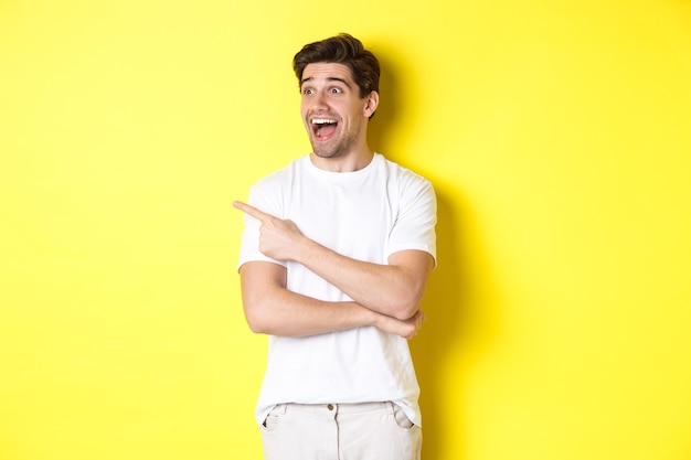 Excité homme souriant pointant et regardant vers la gauche, vérifiant l'offre promotionnelle, debout sur fond jaune.