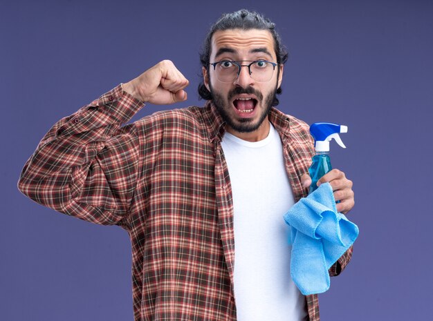 Excité jeune beau mec de nettoyage portant t-shirt tenant un flacon pulvérisateur avec un chiffon montrant oui geste isolé sur mur bleu