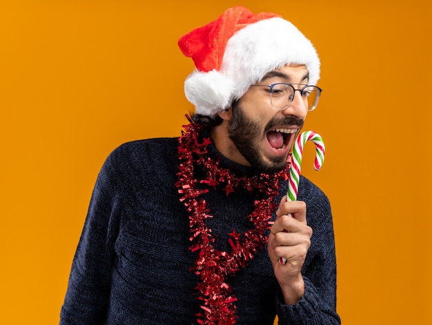 Excité jeune beau mec portant un chapeau de Noël avec guirlande sur le cou tenant des bonbons de Noël isolé sur fond orange