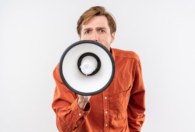 Excité jeune beau mec vêtu d'une chemise rouge parle sur haut-parleur isolé sur mur blanc