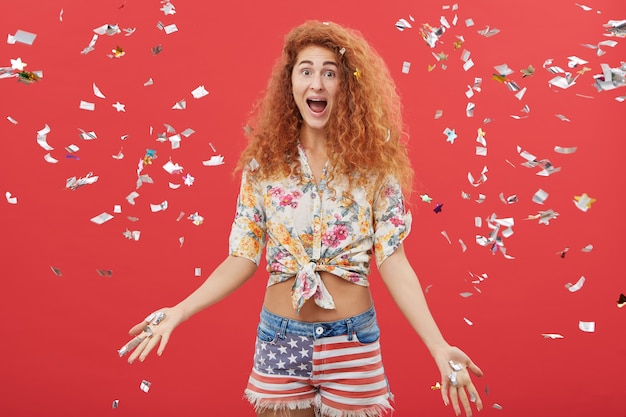 Excité jeune femme jetant des papiers en l'air en se tenant debout sur fond de studio rouge