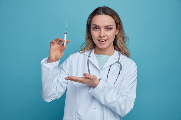 Excité jeune femme médecin portant une robe médicale et un stéthoscope autour du cou tenant et pointant avec la main à la seringue