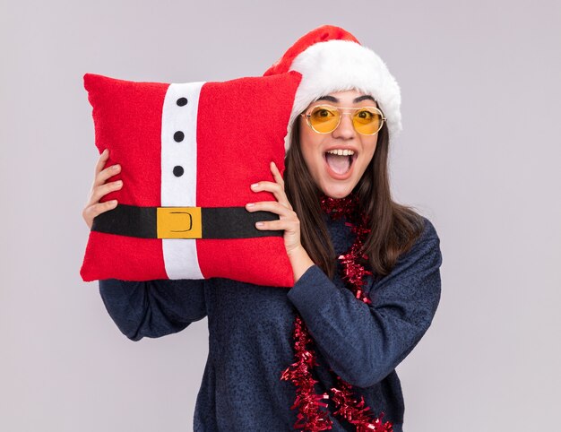 Excité jeune fille de race blanche avec Bonnet de Noel et guirlande autour du cou détient oreiller décoré isolé sur fond blanc avec espace copie