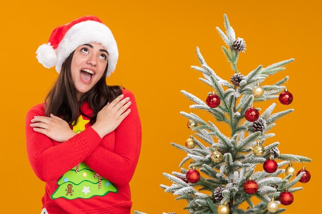 Excité jeune fille de race blanche avec Bonnet de Noel met les mains sur les épaules en levant debout à côté de l'arbre de Noël isolé sur fond orange avec copie espace