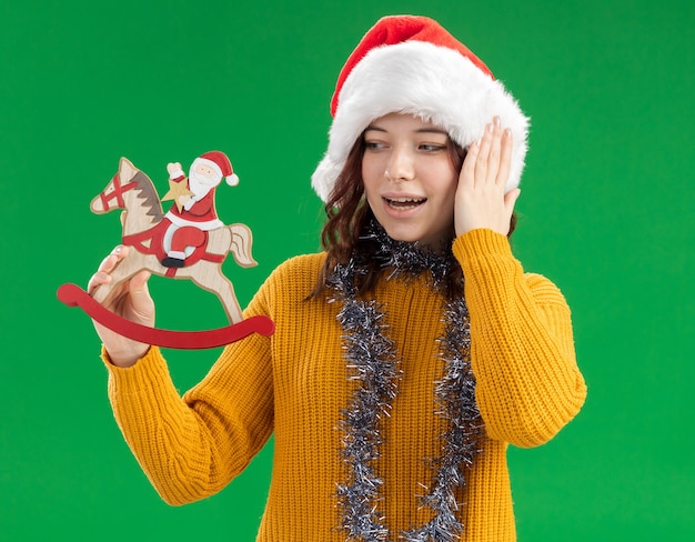 Excité jeune fille slave avec bonnet de Noel et avec guirlande autour du cou tenant et regardant le père Noël sur la décoration de cheval à bascule