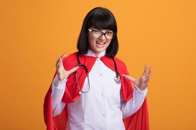 Excité jeune fille de super-héros portant un stéthoscope avec une robe médicale et une cape avec des lunettes écartant les mains