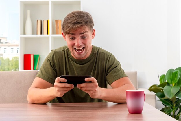 Excité jeune homme beau blond est assis à table avec tasse tenant et regardant le téléphone à l'intérieur du salon