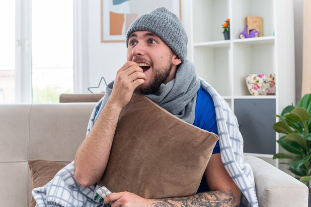 Excité jeune homme malade portant une écharpe et un chapeau d'hiver enveloppé dans une couverture assis sur un canapé dans le salon tenant un oreiller en gardant la main sur la bouche en regardant de côté