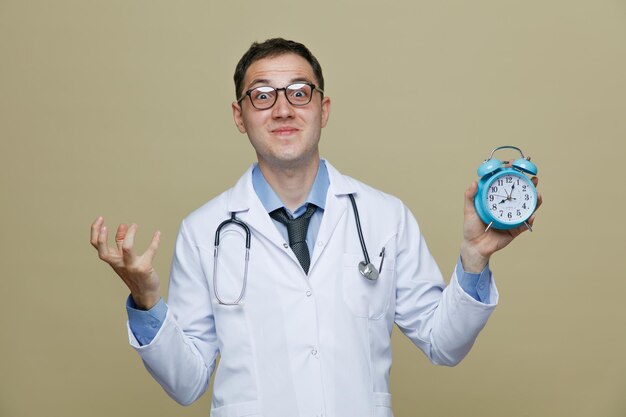 Excité jeune homme médecin portant des lunettes robe médicale et stéthoscope autour du cou regardant la caméra montrant réveil et main vide isolé sur fond vert olive