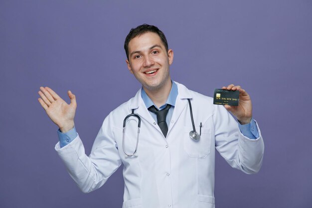 Excité jeune homme médecin portant une robe médicale et un stéthoscope autour du cou regardant la caméra montrant la carte de crédit et la main vide isolé sur fond violet