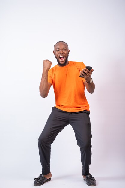 Excité jeune homme nigérian célèbre tout en regardant son téléphone isolé sur un mur blanc