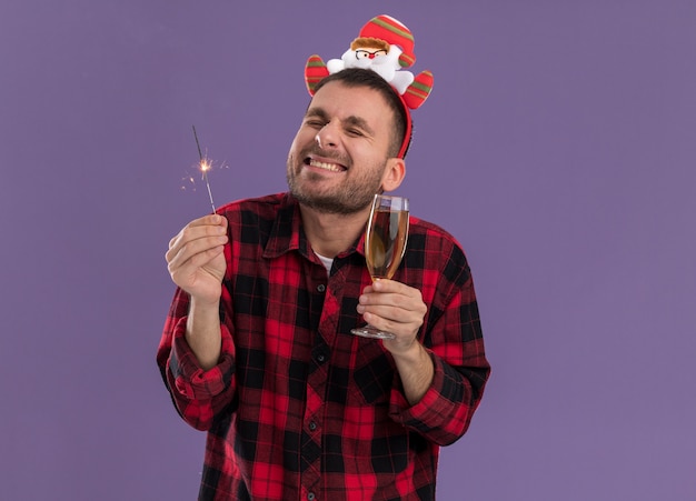 Excité jeune homme de race blanche portant le bandeau du père noël tenant un cierge de vacances et un verre de champagne souriant avec les yeux fermés isolé sur fond violet