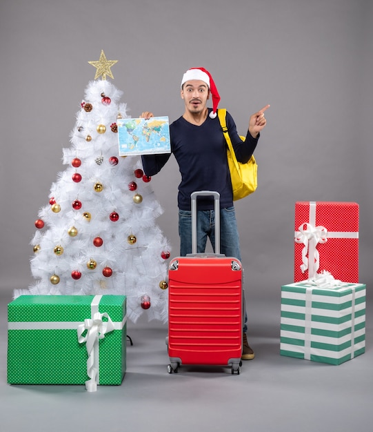 Excité jeune homme avec sac à dos jaune tenant la carte près de l'arbre de Noël blanc sur fond gris