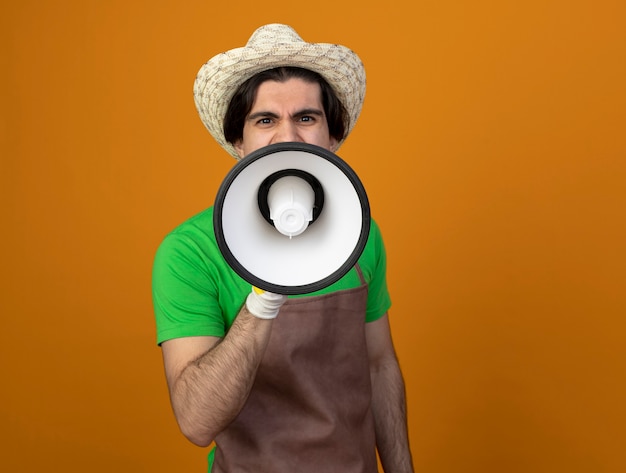 Excité jeune jardinier mâle en uniforme portant chapeau de jardinage avec des gants parle sur haut-parleur