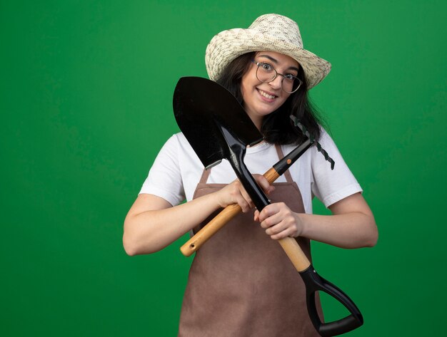 Photo gratuite excité jeune jardinière brune à lunettes optiques et uniforme portant chapeau de jardinage détient pelle et râteau isolé sur mur vert