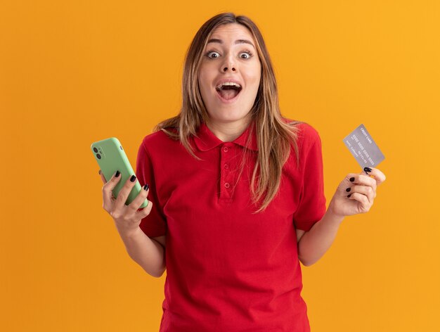 Excité jeune jolie fille caucasienne détient carte de crédit et téléphone sur orange