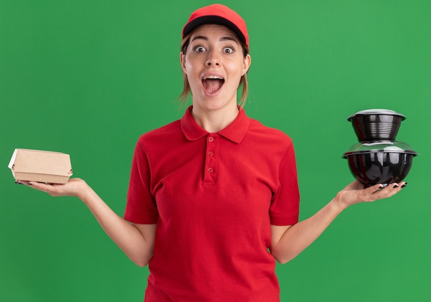 Excité jeune jolie livreuse en uniforme détient des récipients et des emballages alimentaires sur vert