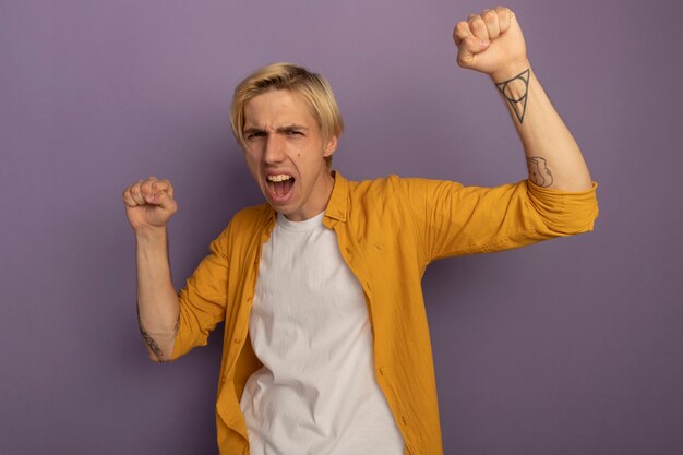 Excité jeune mec blond portant un t-shirt jaune, levant les poings isolés sur violet