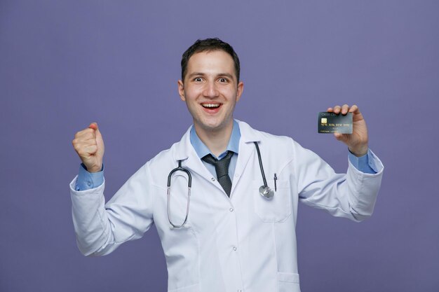 Excité jeune médecin de sexe masculin portant une robe médicale et un stéthoscope autour du cou regardant la caméra montrant la carte de crédit et le poing isolé sur fond violet