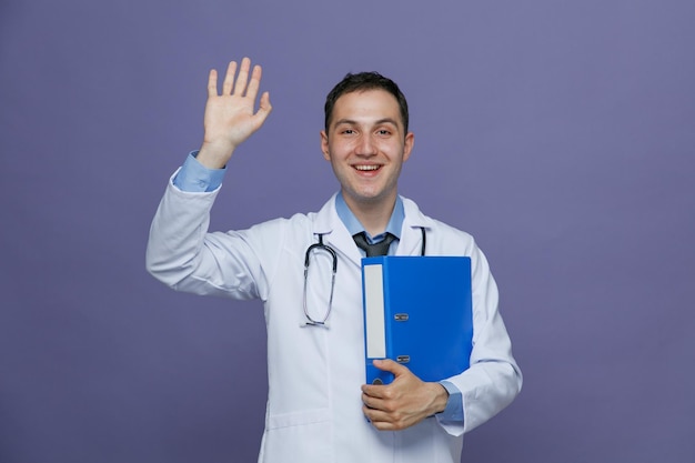 Excité jeune médecin de sexe masculin portant une robe médicale et un stéthoscope autour du cou tenant un dossier le serrant en regardant la caméra montrant le geste salut isolé sur fond violet