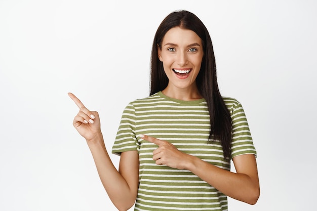 Excité jolie femme pointant les doigts vers la gauche souriant et montrant la publicité de remise de vente en magasin debout sur fond blanc