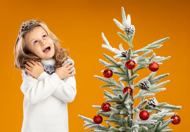 Excité jusqu'à la petite fille debout à proximité de l'arbre de Noël portant tiare avec guirlande sur le cou mettant la main sur les épaules isolé sur mur orange