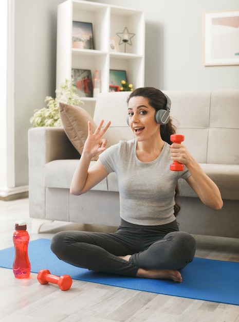 Excité montrant un geste correct jeune fille portant des écouteurs faisant de l'exercice avec des haltères sur un tapis de yoga devant un canapé dans le salon