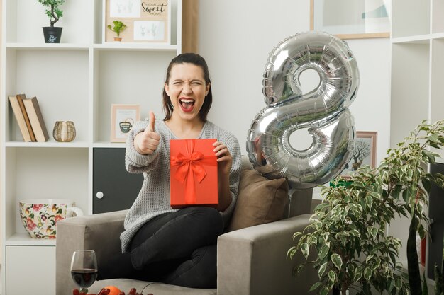 Excité montrant le pouce vers le haut belle femme le jour de la femme heureuse tenant présent assis sur un fauteuil dans le salon