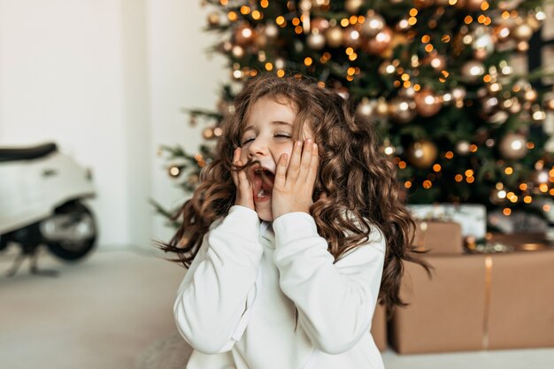 Excité petite fille avec des émotions surprises couvrant le visage avec les mains assis en face de l'arbre de Noël et en attente de cadeaux de Noël