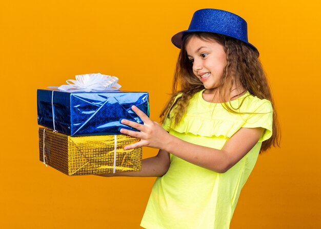 Excité petite fille de race blanche avec chapeau de fête bleu tenant et regardant les coffrets cadeaux isolés sur mur orange avec espace copie