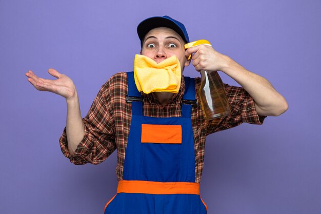 Excité de propagation de la main jeune homme de nettoyage portant un uniforme et une casquette mettant un chiffon dans la bouche tenant un agent de nettoyage sur la tête