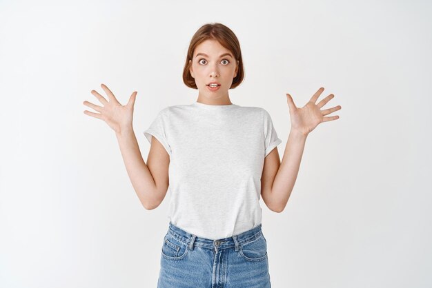 Excité et surpris jeune femme levant les mains et l'air étonné d'expliquer les grandes nouvelles debout en t-shirt et jeans sur fond blanc
