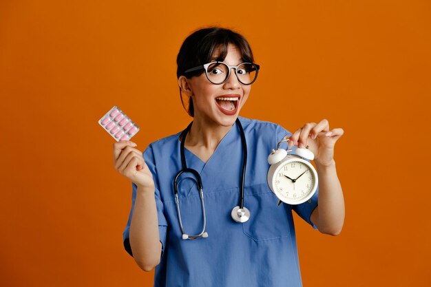 Excité tenant un réveil avec des pilules jeune femme médecin portant un stéthoscope fith uniforme isolé sur fond orange