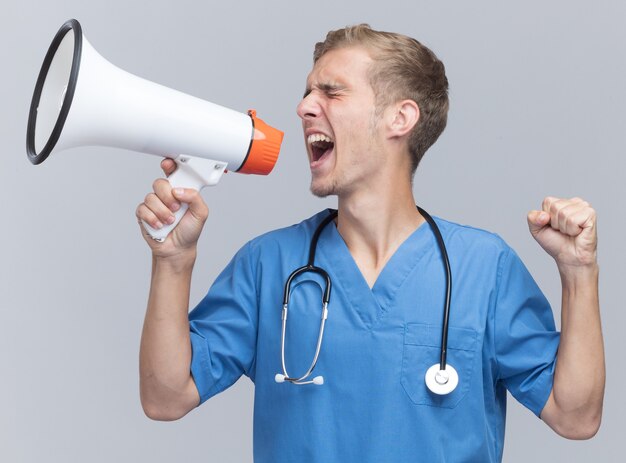 Excité les yeux fermés jeune médecin de sexe masculin portant l'uniforme de médecin avec stéthoscope parle sur haut-parleur montrant oui geste isolé sur mur blanc