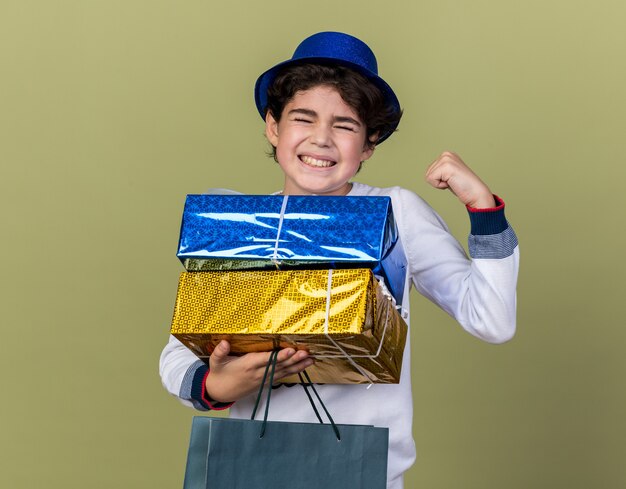 Excité avec les yeux fermés petit garçon portant un chapeau de fête bleu tenant des coffrets cadeaux avec un sac montrant un geste oui isolé sur un mur vert olive