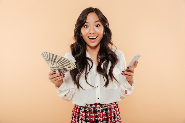 Excited lady holding fan de billets de 100 dollars dans une main et smartphone à la mode dans un autre étant choqué par une énorme somme d'argent sur fond de pêche