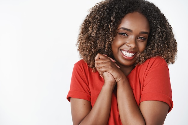 Excitée et accablée mignonne petite amie à la peau foncée amusée en t-shirt rouge souriant largement en appuyant les paumes l'une contre l'autre près de la poitrine, debout, ravie et reconnaissante pour un cadeau génial sur un mur blanc