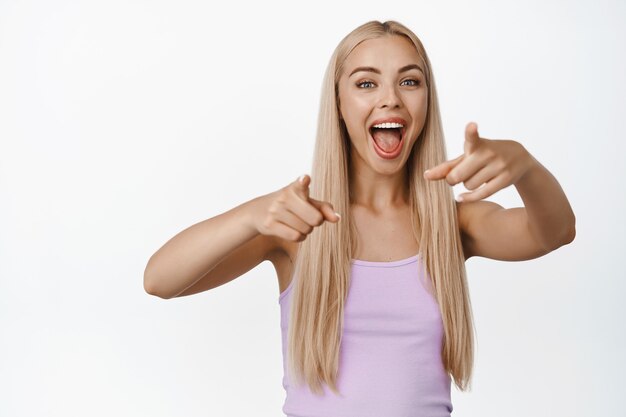 Excitée belle femme pointant du doigt, riant et souriant sur quelque chose de génial, debout dans un débardeur sur blanc.