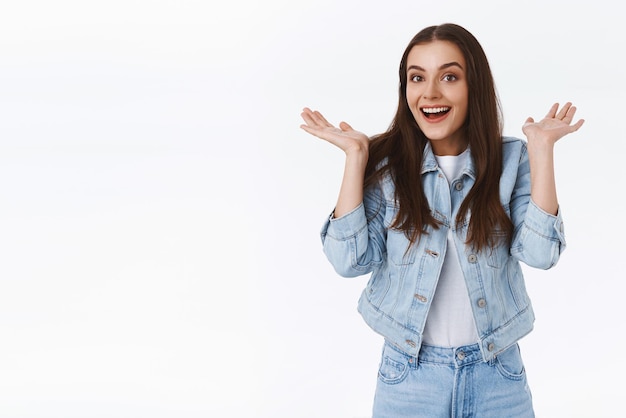 Excitée enthousiaste séduisante femme brune en jeans veste en jean levant les mains surprise et étonnée d'entendre de merveilleuses nouvelles répandre les mains sur le côté impressionné souriant profiter d'un événement génial