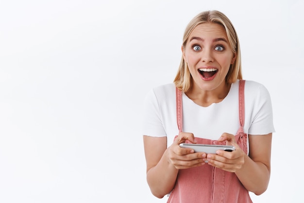 Excitée et heureuse surprise, une jolie fille blonde passe le niveau de jeu difficile et regarde la caméra avec enthousiasme et joie, vous disant qu'elle a gagné, tenant un smartphone horizontalement, debout sur fond blanc