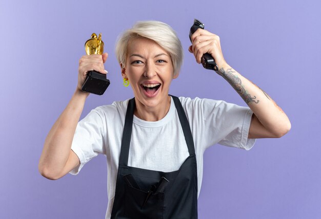 Excitée jeune belle femme barbier en uniforme tenant la coupe du vainqueur avec une tondeuse à cheveux isolée sur un mur bleu