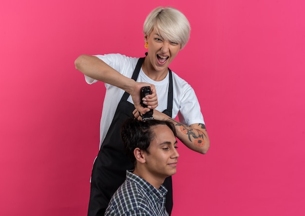 Excitée jeune belle femme barbier en uniforme tenant des outils de barbier et faisant une coupe de cheveux pour un garçon isolé sur un mur rose