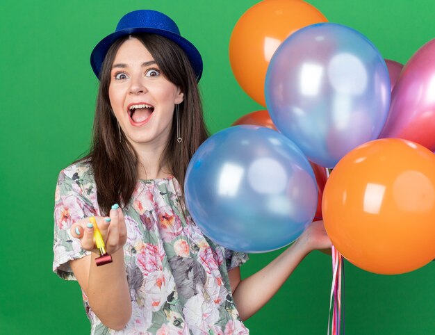 Excitée jeune belle femme portant un chapeau de fête tenant des ballons avec un sifflet de fête isolé sur un mur vert