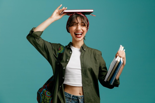 excitée jeune étudiante fille portant bandana et sac à dos tenant des blocs-notes regardant la caméra mettant l'un des blocs-notes sur la tête isolé sur fond bleu